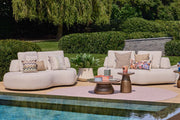 Outdoor patio furniture set arranged around a swimming pool in a landscaped garden. The set includes curved cream boucle lounge chairs, patterned accent pillows, two matching armchairs with throw blankets, and small round coffee tables.
